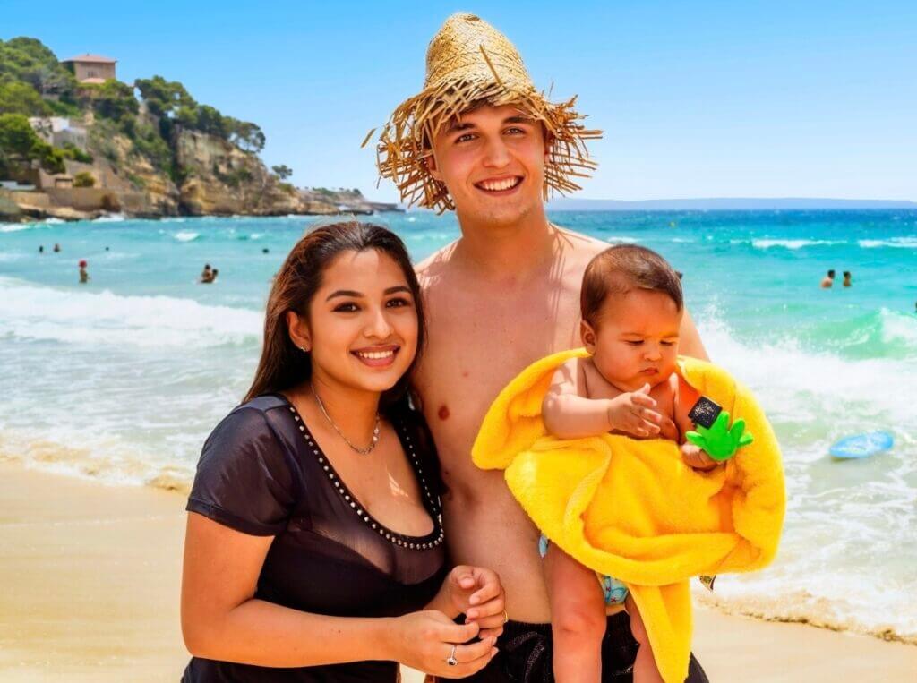 famille-plage Une famille souriante avec un bébé sur une plage ensoleillée avec la mer en arrière-plan.